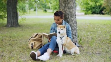 Oldukça Afro-Amerikan kız öğrenci iken iyi yetişmiş köpeğini yanına oturmuş ve hava kokulu ve etrafa park'ın bahçesinde oturup kitap okuma.
