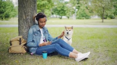Modern Afro-Amerikan Kız okşayarak ve hayvan sevecen ve şehir parkı ile evde beslenen hayvan köpek, smartphone rahatlatıcı kullanarak bir kulaklık ile müzik dinlediği.
