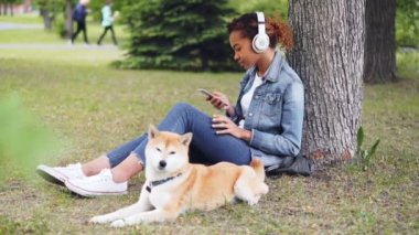 Çekici Afrikalı-Amerikalı kadın kulaklık ile müzik dinlemek ve onu köpek çim yeme yakın yatarken çim parkta oturan smartphone kullanarak.