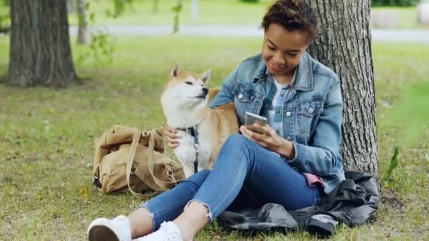 Gezellig meisje is met behulp van smartphone SMS vrienden en streelde haar schattige puppy terwijl rusten in park in weekend. Moderne technologie, mensen en dieren concept. — Stockvideo