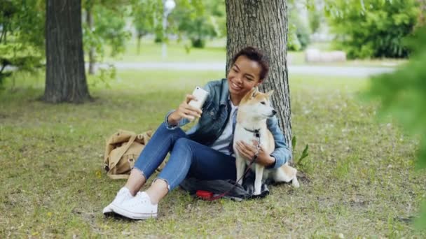 Досить молода дівчина-блогер бере селфі з чистою собакою на відкритому повітрі в міському парку, прикрашаючи і люблячи красиву тварину. Сучасні технології, люблячі тварини та концепція природи . — стокове відео
