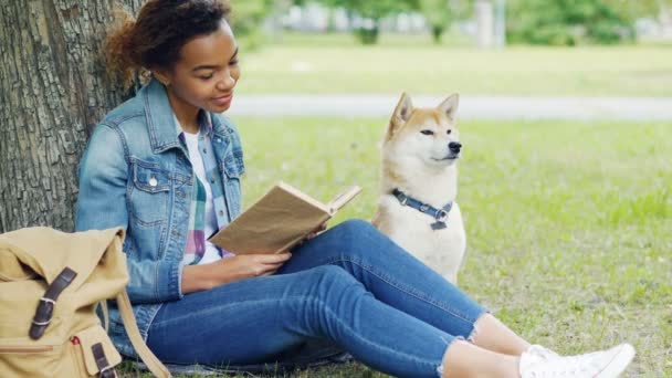 Mujer de raza mixta amable está leyendo libro en el parque y acariciando a su perro sentado en el césped bajo el árbol juntos. Pasatiempo inteligente, acariciar animales y concepto de juventud . — Vídeos de Stock