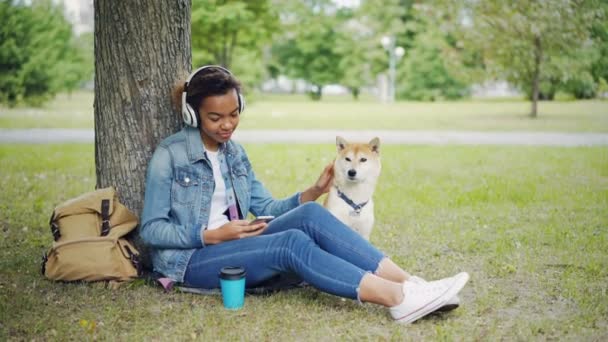 Chica afroamericana moderna está escuchando música con auriculares y el uso de teléfono inteligente relajante en el parque de la ciudad con perro mascota, acariciando y acariciando al animal . — Vídeos de Stock