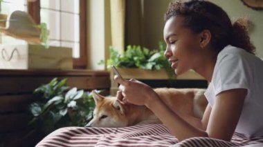 Güzel kız öğrenci ekran dokunaklı ve Sevimli evcil köpek ile evde yatakta yatarken gülüyor smartphone kullanarak, hayvan bakım ve sevgi keyif alıyor.