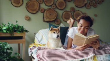 Neşeli karışık yarış genç kız edebiyat zevk sonra yatağın yanında yatan shiba Inu köpeğini okşayarak kitap okuyor. Hobi, modern yaşam tarzı ve hayvanlar kavramı.