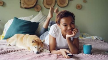 Afrikalı-Amerikalı kadın uzaktan kumanda acil düğmeleri tutarak ve onu evde beslenen hayvan köpek onun yanında yatarken gülüyor televizyon izliyor. Eğlence ve teknoloji kavramı.
