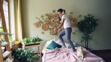 Neşeli stydent kablosuz kulaklık ile müzik dinlemek ve şarkı dans ve sevimli safkan köpek yatakta istirahat ile yatak odasında atlama saç kurutma makinesi olduğunu.