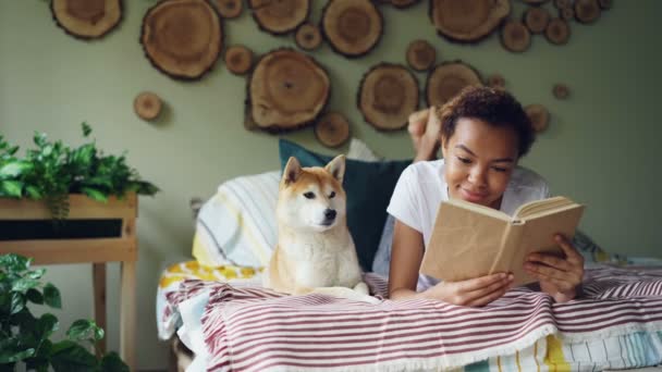 Lachende African American student lief meisje is boek op bed thuis lezen, terwijl haar huisdier hond in de buurt van haar ligt. Hobby, vrije tijd, dieren en interieur concept. — Stockvideo