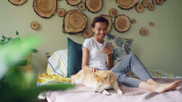 Mujer joven y bonita está utilizando el teléfono inteligente y acariciando cachorro encantador acostado en la cama cerca de su propietario en el dormitorio de estilo moderno en casa. Concepto de tecnología, animales y personas . — Vídeo de stock