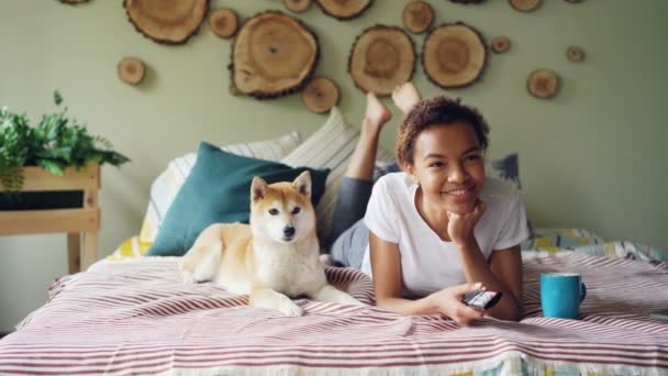 Murid ras campuran yang ceria sedang menonton TV memegang remote dan menekan tombol-tombol memilih saluran televisi sementara anjingnya sedang bergerak di tempat tidur di rumah . — Stok Video