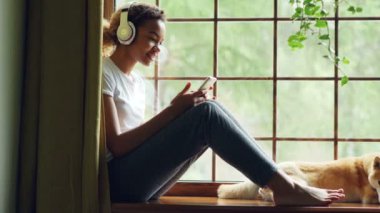 Oldukça Afrikalı-Amerikalı kız ile güzel iyi yetişmiş köpek ona yalan pencere pervazına üzerinde oturan smartphone kullanarak ve kulaklık ile müzik dinlemek.