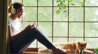 Çekici genç kadın kablosuz kulaklık aracılığıyla müzik dinlemek ve şarkı Smartphone cep telefonu ile seçerek, onu evde beslenen hayvan köpek sahibi yakın pencere yatıyor.