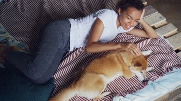 Rodzaju African American Dziewczynka głaszcze, piękny pies, leżąc na łóżku w domu razem, korzystających spokoju i odpoczynku. Nowoczesne drewniane łóżko i jasny len jest widoczny. — Wideo stockowe