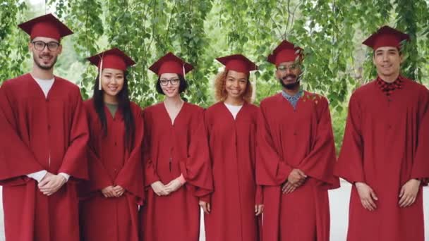 Porträtt av multietnisk grupp examen studenter står utomhus bär röda kappor och murbruk-styrelser, leende och tittar på kameran. — Stockvideo