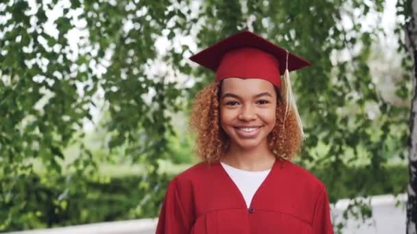 Portret van vrij African American girl afstuderen student in rode jurk en mortarboard permanent buitenshuis, glimlachend en camera kijken. Jeugd en onderwijs concept. — Stockvideo