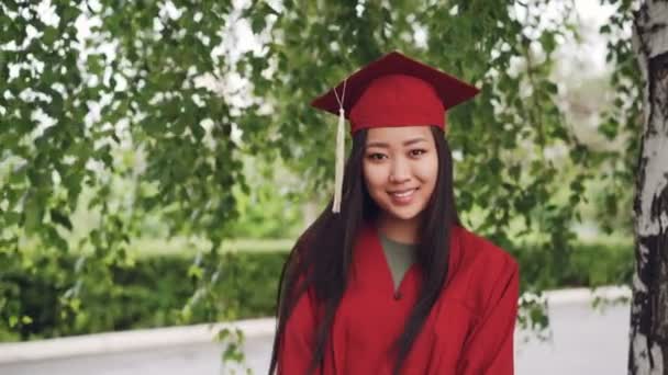 Porträt einer attraktiven asiatischen Studentin, die in Kleid und Pappe auf dem Campus steht, lächelt und in die Kamera blickt. Jugend- und Bildungskonzept. — Stockvideo