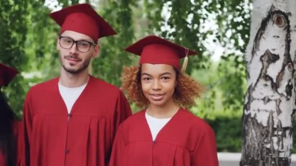 Retrato de grupo multinacional de estudantes graduados em vestidos de graduação vermelho e placas de argamassa em pé juntos ao ar livre, sorrindo e olhando para a câmera . — Vídeo de Stock