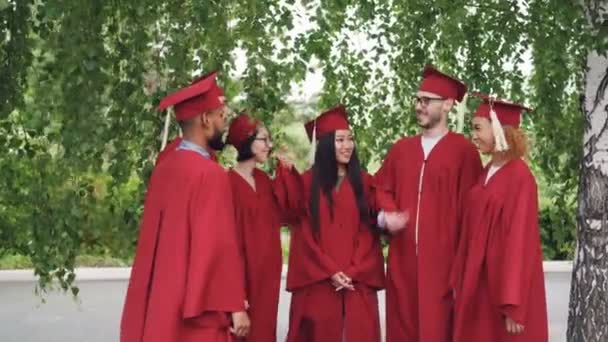 Joyeux diplômés groupe multiracial étudiants mettent la main ensemble puis applaudissent les mains célébrant la graduation, amis sont debout à l'extérieur portant des robes et des mortiers-panneaux . — Video