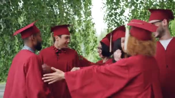 Opgewonden jongeren afstuderen studenten in jassen en hoeden zijn knuffelen elkaar feliciteren met afstuderen, lachen en vieren einde van academisch jaar. — Stockvideo