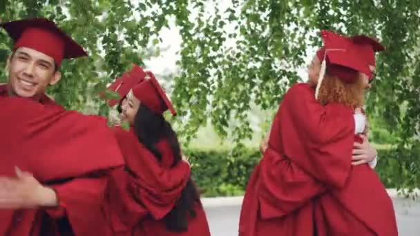 Amigos alegres graduando-se estudantes em vestidos e almofadas estão se parabenizando após a cerimônia de formatura, eles estão abraçando e rindo . — Vídeo de Stock
