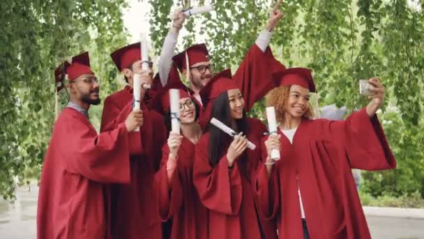 Los estudiantes graduados emocionados están tomando selfie con el teléfono inteligente, los jóvenes están saludando diplomas, posando, sonriendo y riendo. Concepto de educación y éxito . — Vídeo de stock