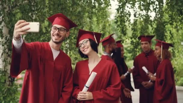 Alegre pareja de estudiantes graduados está tomando selfie usando teléfono inteligente, hombre joven y mujer están sosteniendo diplomas, mirando a la cámara del teléfono inteligente y sonriendo . — Vídeo de stock