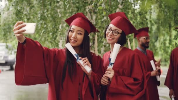 Chicas guapas graduándose estudiantes están tomando selfie con pergaminos de diploma usando teléfono inteligente, las mujeres están posando y sonriendo, sus compañeros estudiantes están celebrando en segundo plano . — Vídeo de stock