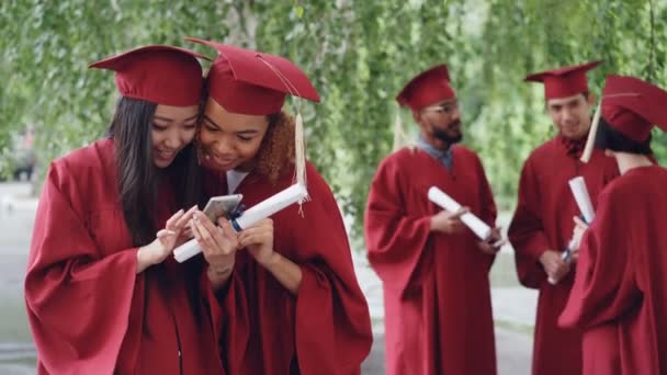 Atractivas mujeres jóvenes graduados felices están viendo fotos en el teléfono inteligente y hablando en el día de la graduación, mientras que sus compañeros de estudios están charlando en segundo plano . — Vídeo de stock