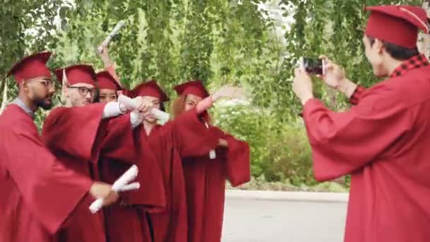 El joven con smartphone está tomando fotos de graduados divirtiéndose posando con diplomas moviendo manos y dedos y gritando con felicidad. Concepto educación y juventud . — Vídeos de Stock