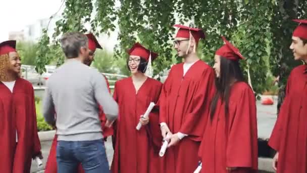 Бородатий чоловік гордий вчитель вітає випускників, які тремтять руками і обіймають їх на відкритому повітрі в кампусі коледжу, поки студенти розмовляють і тримають дипломи . — стокове відео
