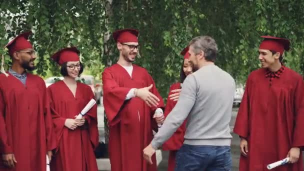 Stolt förälder gratulera studenter examen dag, skakar hand och kramar dem på bakgrund av universitetsområdet, unga människor pratar och ler. — Stockvideo