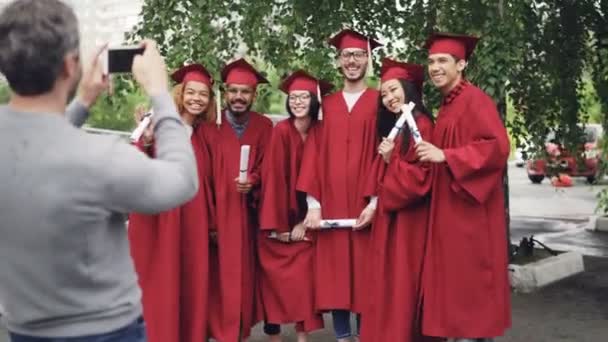 Stolt far är att ta bilder av examen studenter med smartphone medan ungdomarna poserar, viftande händer med diplom och gestikulerande. Teknik och utbildning koncept. — Stockvideo