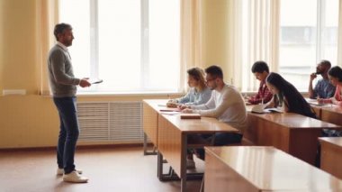 Rahat kıyafetler içinde erkek Profesör öğrencilerin sınıfta masalarda oturan ve notlar grubuna konuşuyor. Masalar, sandalyeler ve windows ile büyük konferans salonunun görülür.