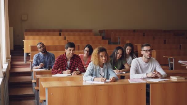 Estudiantes diligentes están escribiendo notas en cuadernos sentados en la conferencia en la universidad mientras el profesor habla de pie frente a ellos en un aula grande . — Vídeo de stock