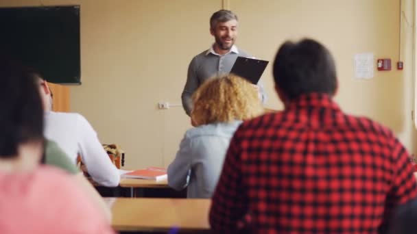 Knappe man van de leraar van de middelbare school is in gesprek met studenten die aan tafels in de klas en glimlachen zitten. Onderwijs, onderwijs- en leerproces, mensen concept. — Stockvideo