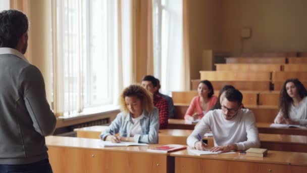 El profesor barbudo está leyendo la conferencia hablando y haciendo gestos mientras los estudiantes escuchan y escriben sentados en las mesas de las amplias aulas universitarias. . — Vídeos de Stock