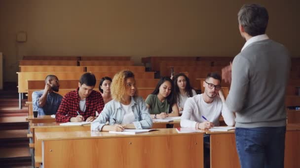 Tutor está hablando con los estudiantes sentados en los escritorios en la espaciosa sala de conferencias de la universidad, los jóvenes con ropa casual están haciendo preguntas. Educación y concepto universitario . — Vídeo de stock