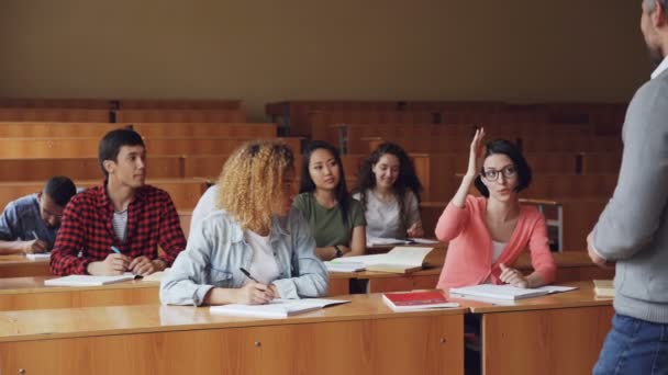 かなり若い女性の勤勉な学生は他の学生の笑顔と聞く中、机に座っている高校教師に話しています。教育および青年の近代的な概念. — ストック動画
