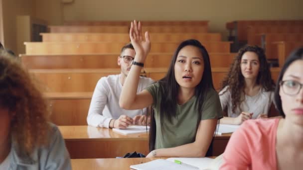 Menina asiática bonita está levantando a mão e fazer perguntas durante a palestra na faculdade, seus colegas de classe estão sentados em mesas sorrindo e rindo. Conceito de educação e juventude . — Vídeo de Stock