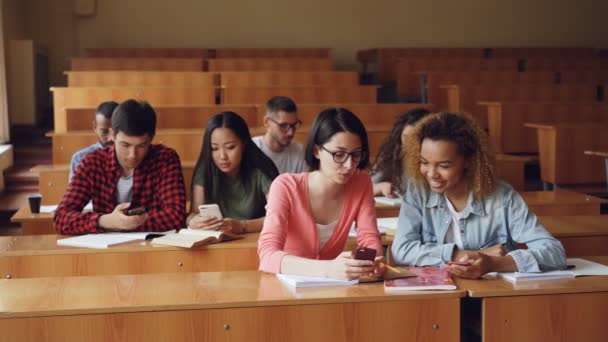 Ungdomar använder smartphones och pratar sitter vid skrivbord i föreläsningssalen på universitet. Rymligt rum med trämöbler, kvinnor och män, anteckningsböcker är synliga. — Stockvideo