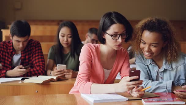 Las niñas bonitas estudiantes están usando teléfonos inteligentes, viendo la pantalla, hablando y riendo sentados en los escritorios de la universidad. Redes sociales, internet, millennials y concepto educativo . — Vídeos de Stock