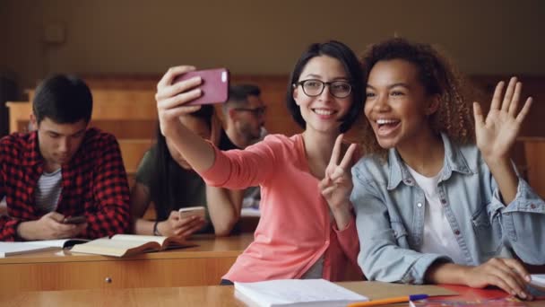 Alunos meninas alegres estão tomando selfie com smartphone sentado em mesas na faculdade, as mulheres estão posando fazendo gestos de mão e abraçando. Conceito de amigos e tecnologia . — Vídeo de Stock