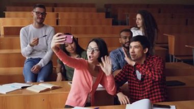 Neşeli öğrenciler kayıt sınıf sallayarak elinde video ve gülümseyen masaları konferans salonunda oturup eğlenceli yaşıyorsanız. Selfie, smartphone ve eğitim kavramı.
