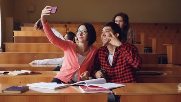 Glada studenter tar selfie i föreläsningssalen sitter tillsammans vid skrivbord och hålla smartphones. Modern teknik, självporträtt och utbildning koncept. — Stockvideo