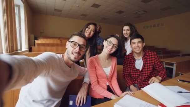 Punto de vista plano de grupo multiétnico de amigos tomando selfie en el aula mirando a la cámara, posando con gestos de mano y sonriendo. Tecnología moderna y concepto de los millennials . — Vídeos de Stock