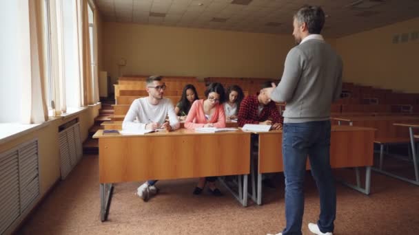Pilní studenti psaní testu sedí u přepážek v přednáškovém sále, zatímco mladý muž v brýlích se vyptávat mluvit s učitelem. Koncept vzdělávání, znalosti a lidé. — Stock video