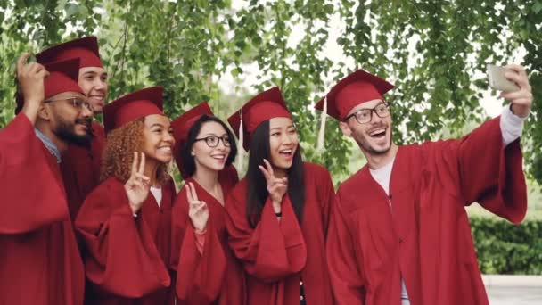 Zwolnionym tempie wesoły studentów graduating biorąc selfie z smartphone, młodzi ludzie są pozowanie, wyświetlone gestów, Śmieszne miny, śmiejąc się. — Wideo stockowe