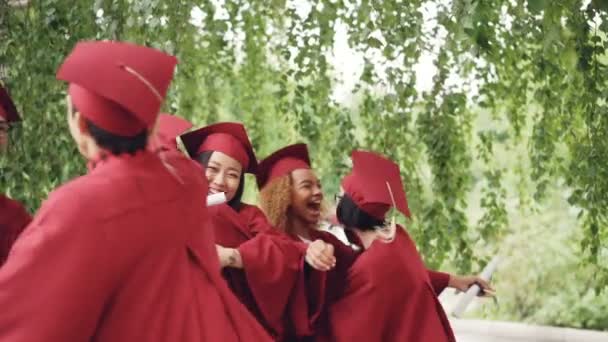 Slow motion van vrolijke mensen elkaar knuffelen na graduatie ceremonie bezit van diploma's en het dragen van de traditionele hoeden en jassen. Opwinding en viering concept. — Stockvideo