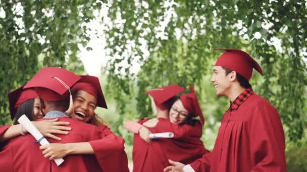 Lycklig examen studenter multietnisk grupp är kramar och gör high-five efter examensceremoni, tjejer och killar i murbruk-styrelser och kappor skratta och ha roligt. — Stockvideo