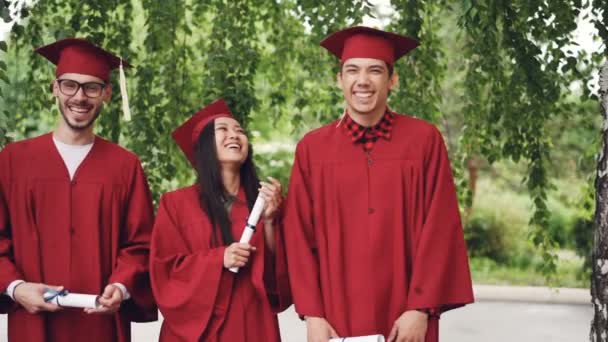 慢动作肖像的毕业生站在排队持有文凭和笑看相机。女孩和男孩穿着迫击炮板和礼服. — 图库视频影像